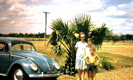 Filmstill zu "Zuhause ist kein Ort" zeigt einen geparkten Käfer (Auto) und eine Mutter mit zwei Kinder, die vor einer Palme posieren. Ein Reise-Motiv 